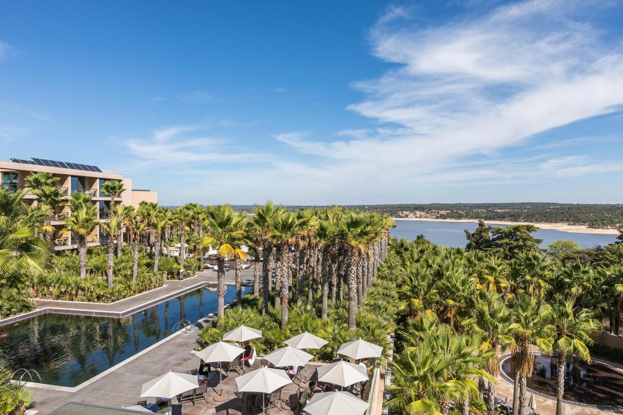 Ap Lago Montargil - Conference & Spa מראה חיצוני תמונה The photo shows a scenic view of a resort or hotel area. In the foreground, there is a pool surrounded by palm trees and lounge chairs with umbrellas, suggesting a relaxing atmosphere. Beyond the pool, there is a body of water, likely a river or coas