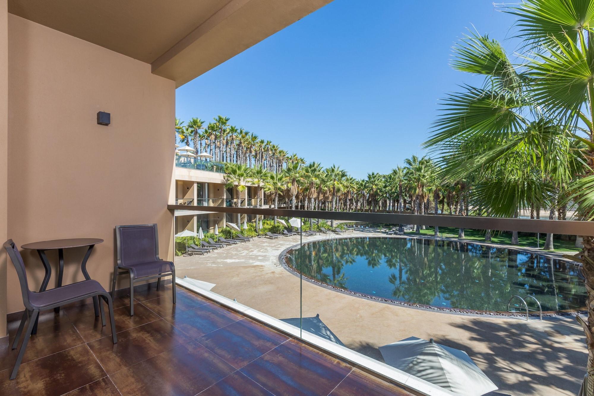 Ap Lago Montargil - Conference & Spa מראה חיצוני תמונה The photo shows a balcony view of a resort area. There is a small round table with two chairs on the balcony, providing a spot to sit and enjoy the view. Below, a large, circular pool is visible, surrounded by palm trees and sun loungers. The sky is 
