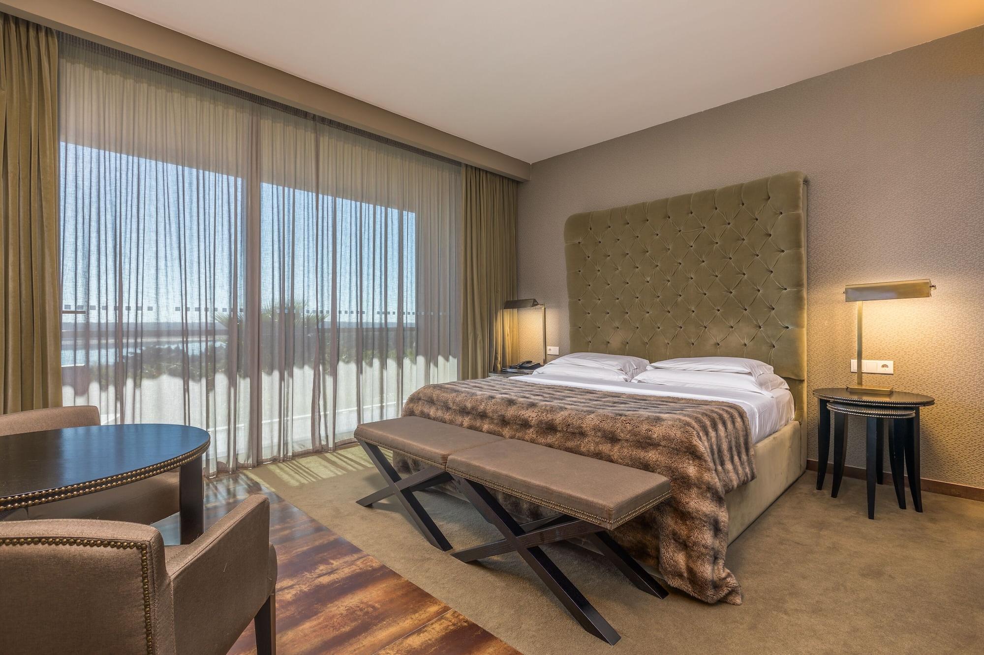 Ap Lago Montargil - Conference & Spa מראה חיצוני תמונה The photo shows a modern hotel room featuring a large bed with a tufted headboard and a cozy blanket. There are curtains draped over large windows that allow natural light to flow in, providing a view of the outside. A small seating area includes two