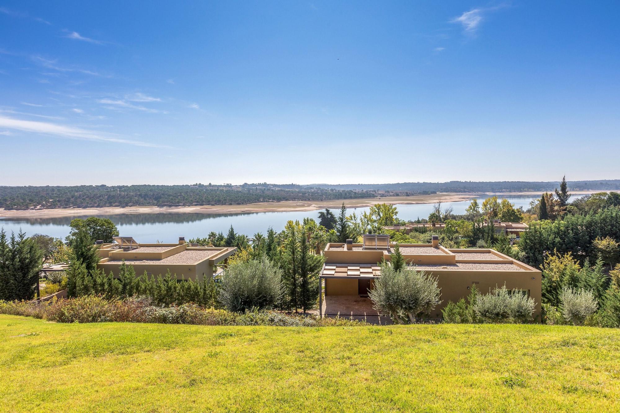 Ap Lago Montargil - Conference & Spa מראה חיצוני תמונה The photo depicts a scenic landscape featuring a calm body of water, possibly a lake or river, surrounded by lush greenery and trees. In the foreground, there are several modern buildings with flat roofs, set against a vibrant blue sky. The overall s