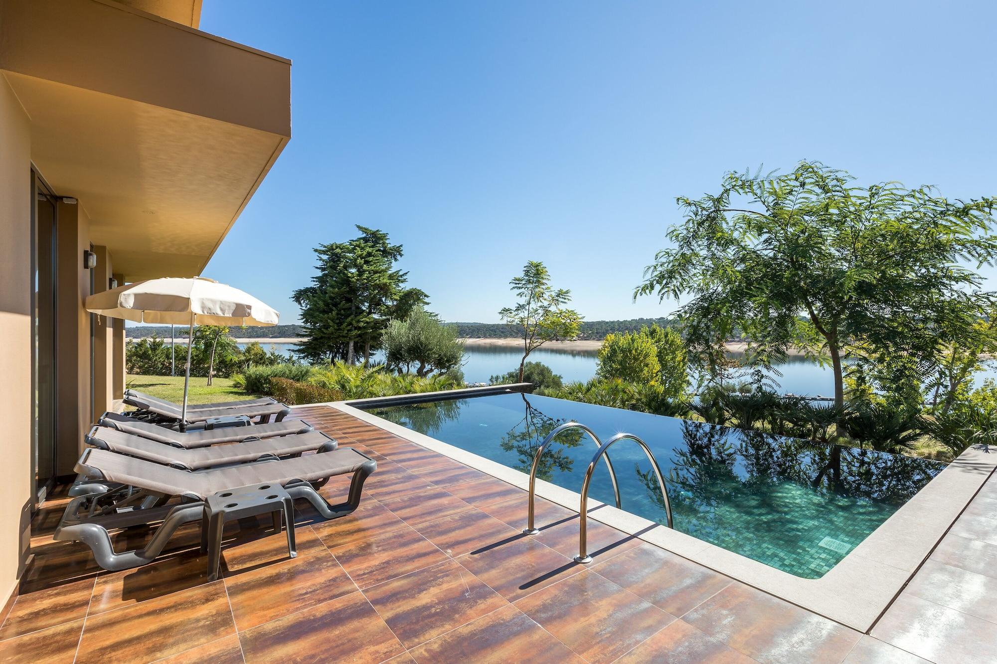 Ap Lago Montargil - Conference & Spa מראה חיצוני תמונה The photo shows a luxurious outdoor setting by a pool. There are several lounge chairs arranged next to an infinity pool, which overlooks a scenic body of water and lush greenery. A parasol provides shade for relaxation. The sky is clear and blue, in