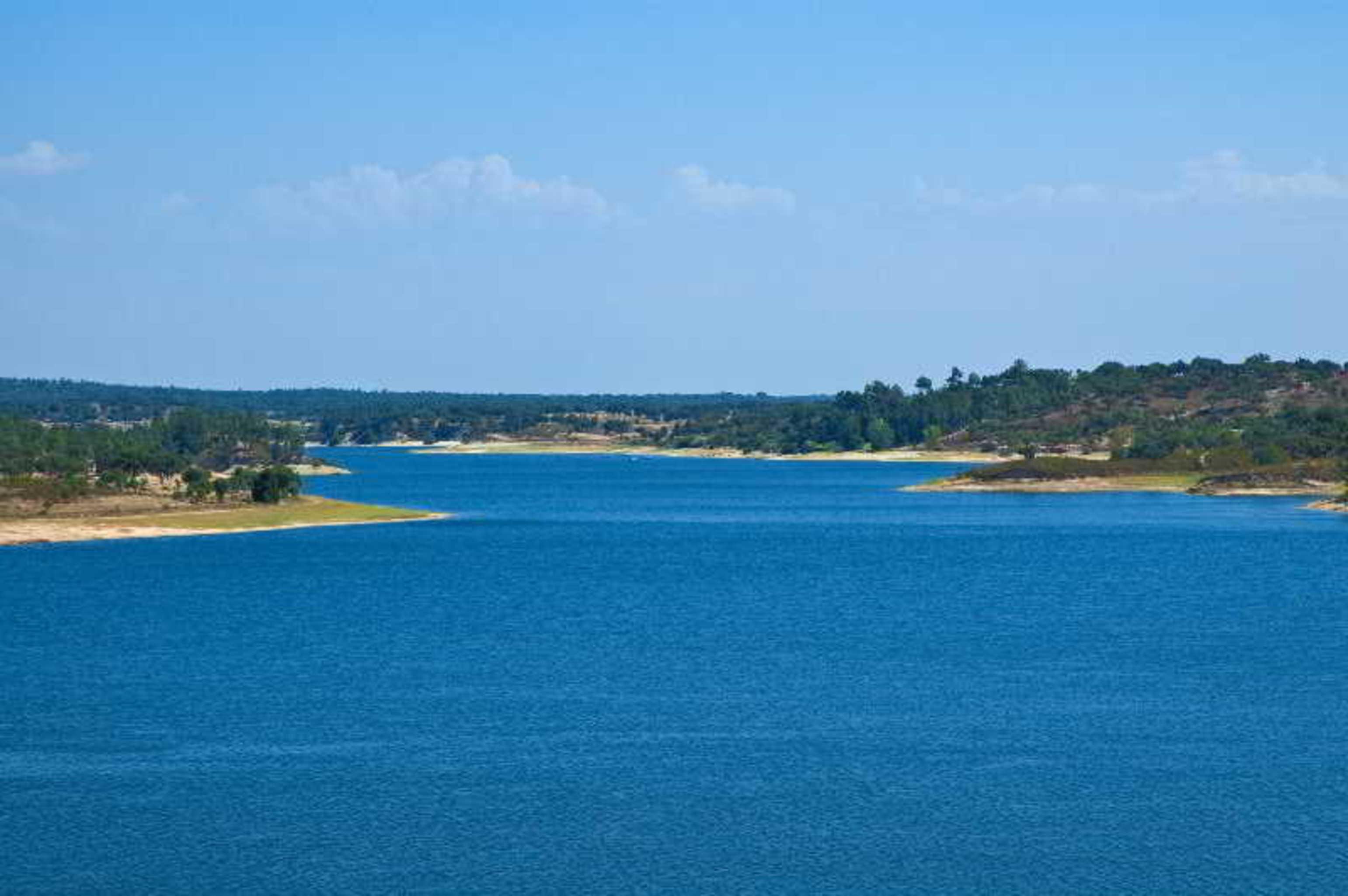 Ap Lago Montargil - Conference & Spa מראה חיצוני תמונה The photo depicts a serene landscape featuring a large body of water, likely a lake or river. The water appears calm and is reflecting a clear blue sky with some clouds. Surrounding the water, there are patches of green land and trees, indicating a n