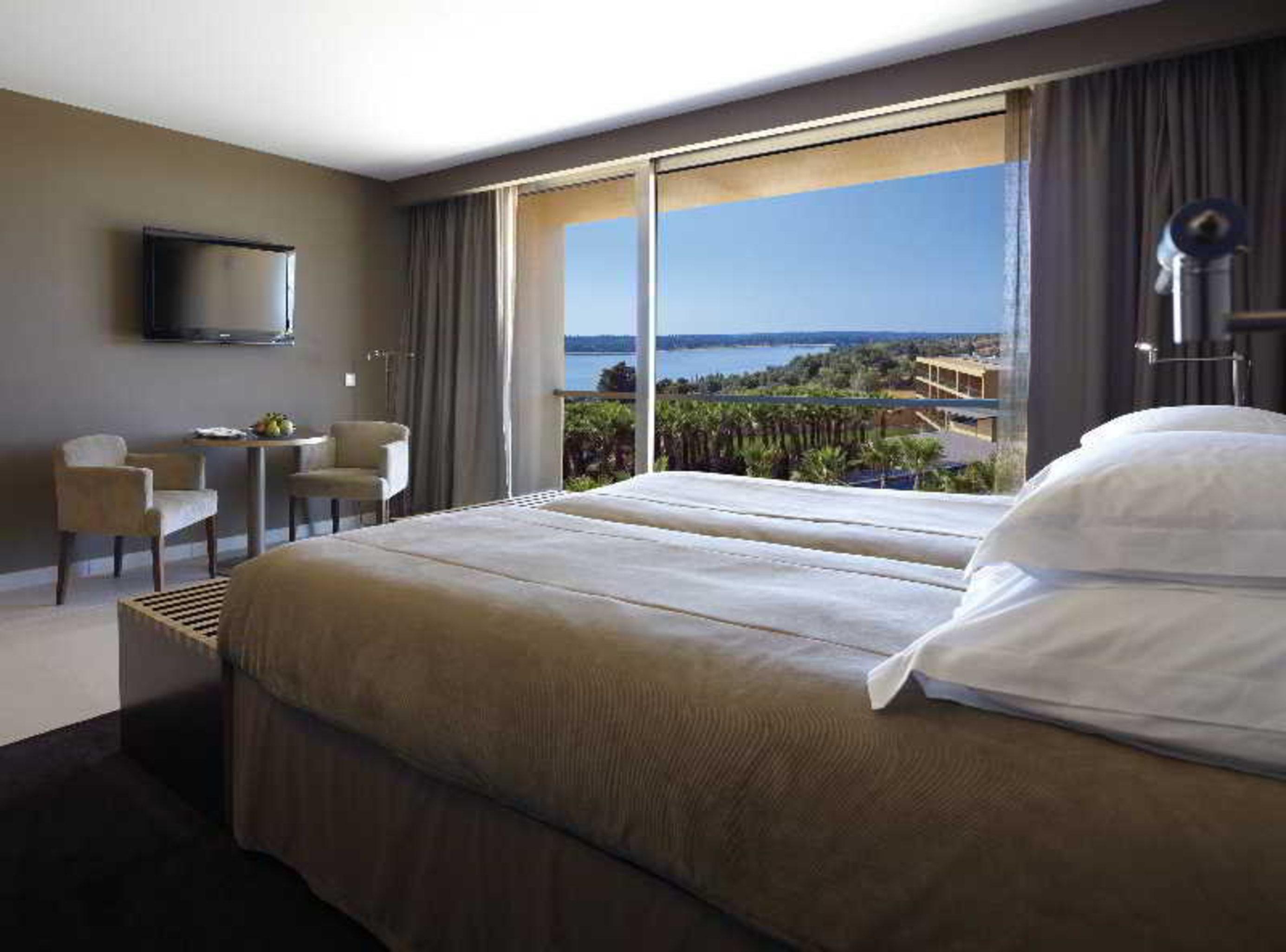 Ap Lago Montargil - Conference & Spa מראה חיצוני תמונה The photo shows a modern hotel room with a large window offering a view of the sea and mountains. The room features a comfortable bed with white pillows and brown bedding. There is a small sitting area with two chairs and a table, as well as a wall-m
