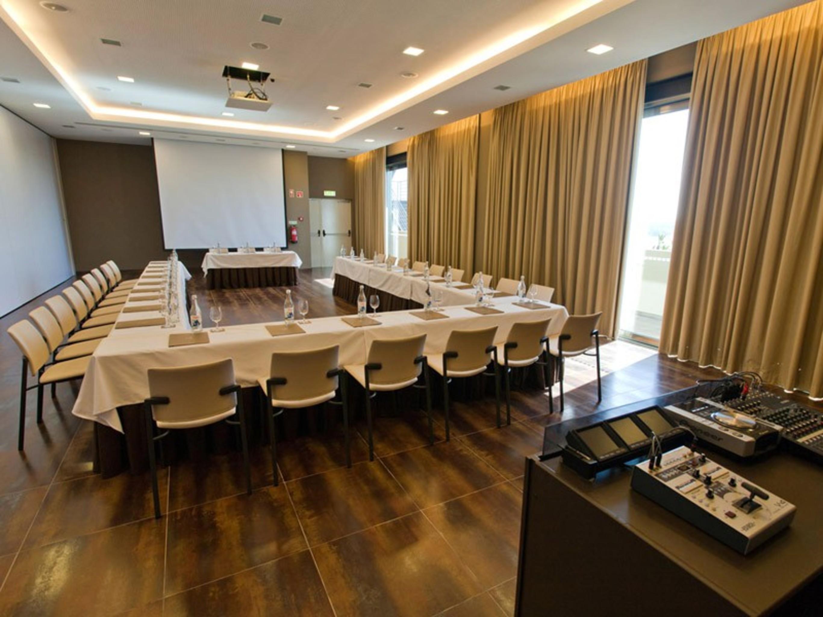 Ap Lago Montargil - Conference & Spa מראה חיצוני תמונה The photo shows a modern conference room set up for a meeting. There are several tables arranged in a U-shape, surrounded by chairs. A large screen and projector are positioned at the front, with a table for speakers or presenters. The room has large