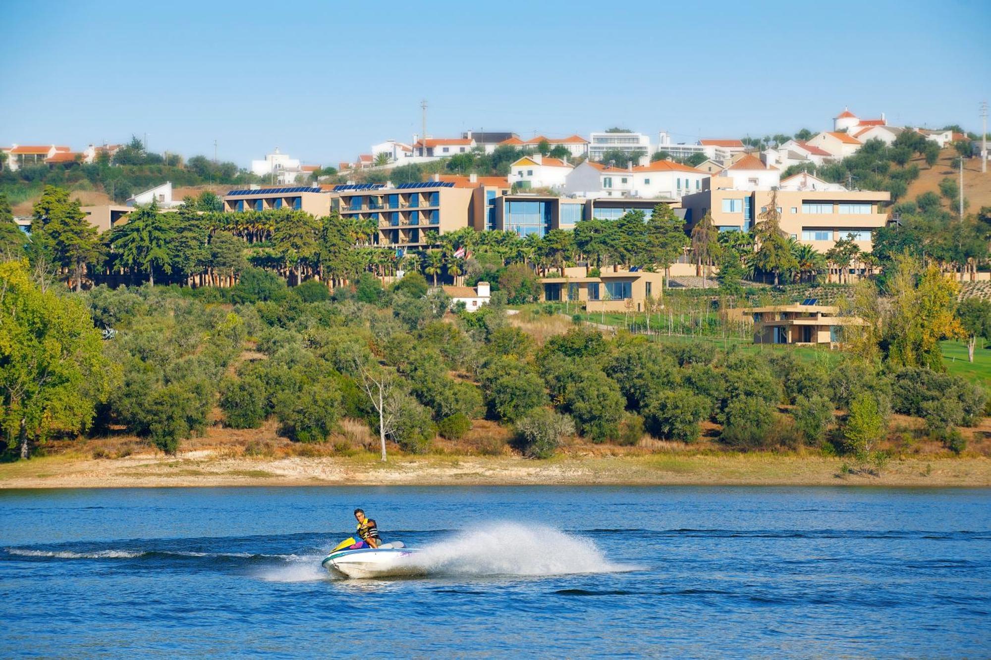 Ap Lago Montargil - Conference & Spa מראה חיצוני תמונה The photo shows a person riding a jet ski on a body of water, creating a spray as they speed across the surface. In the background, there are modern buildings and a hillside with trees and greenery. The sky is clear and blue, suggesting a bright and 